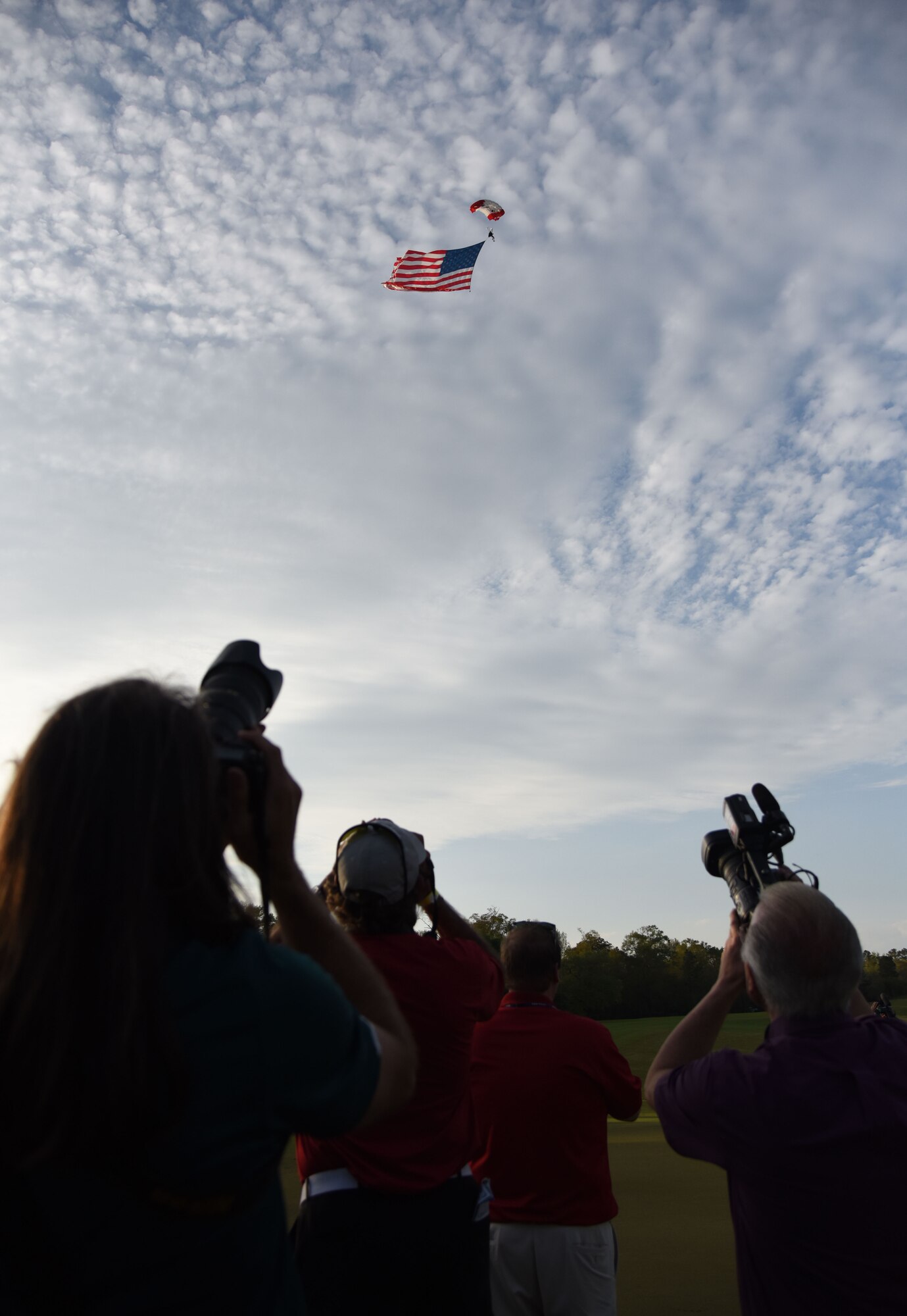 News cameras document retired U.S. Army Sgt. 1st Class Dana Bowman, a double amputee, parachute down while carrying the U.S. flag during the Rapiscan Systems Classic Champions Tour at Fallen Oak Golf Club March 25, 2018, in Saucier, Mississippi. Keesler personnel performed the national anthem and presented the colors during the closing ceremony of the three-day event. This is the fifth year that the event has been held on the coast. (U.S. Air Force photo by Kemberly Groue)