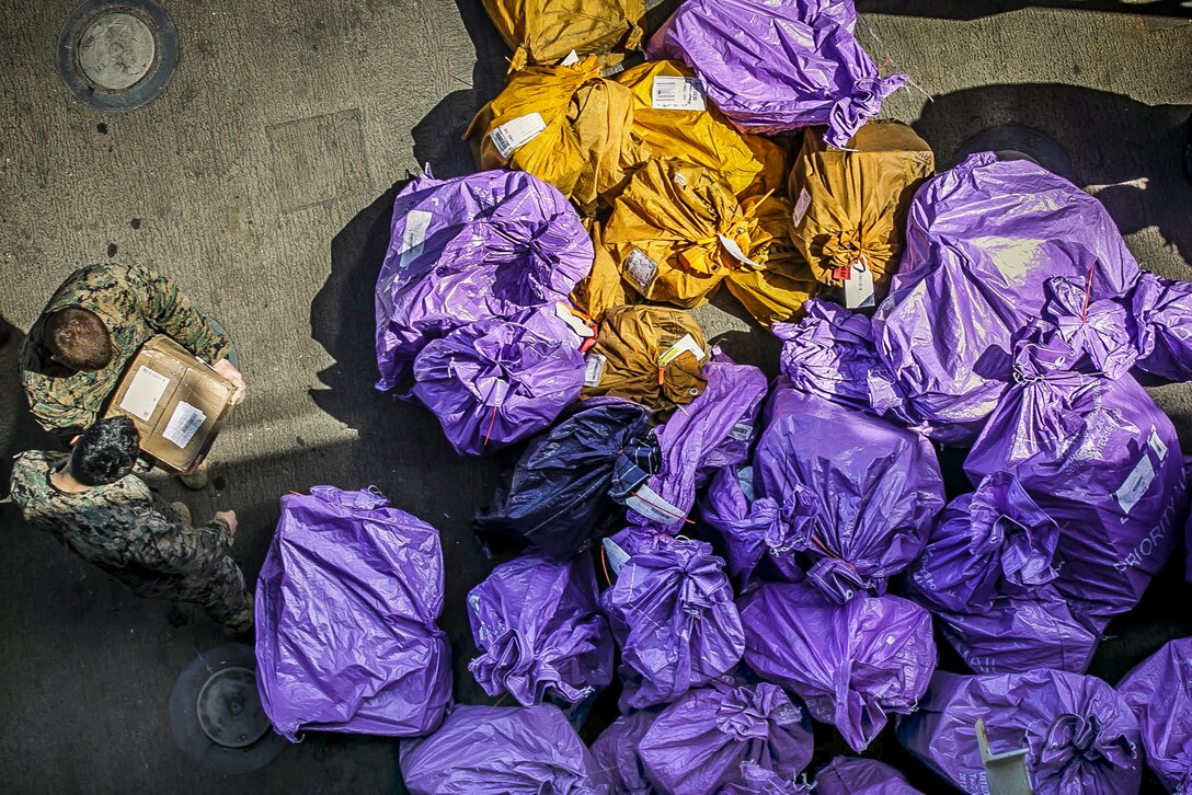 Two Marines, shown from overhead, hold a package while standing by big purple bags of mail.