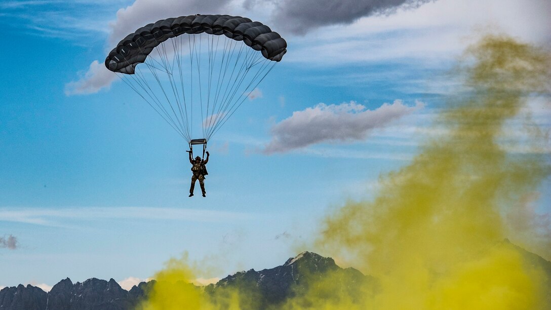 An airman parachutes over mountain peaks, as yellow smoke wafts nearby.