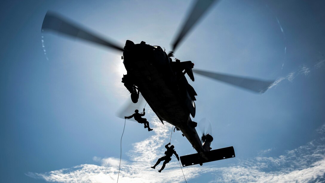 Two sailors, shown in silhouette, rappel out of a helicopter on ropes.