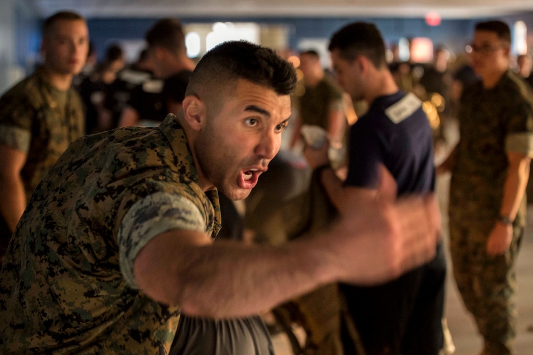 A Marine waves his arm and yells in a room where other Marines are milling about.