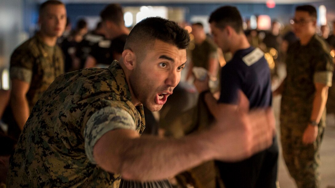 A Marine waves his arm and yells in a room where other Marines are milling about.