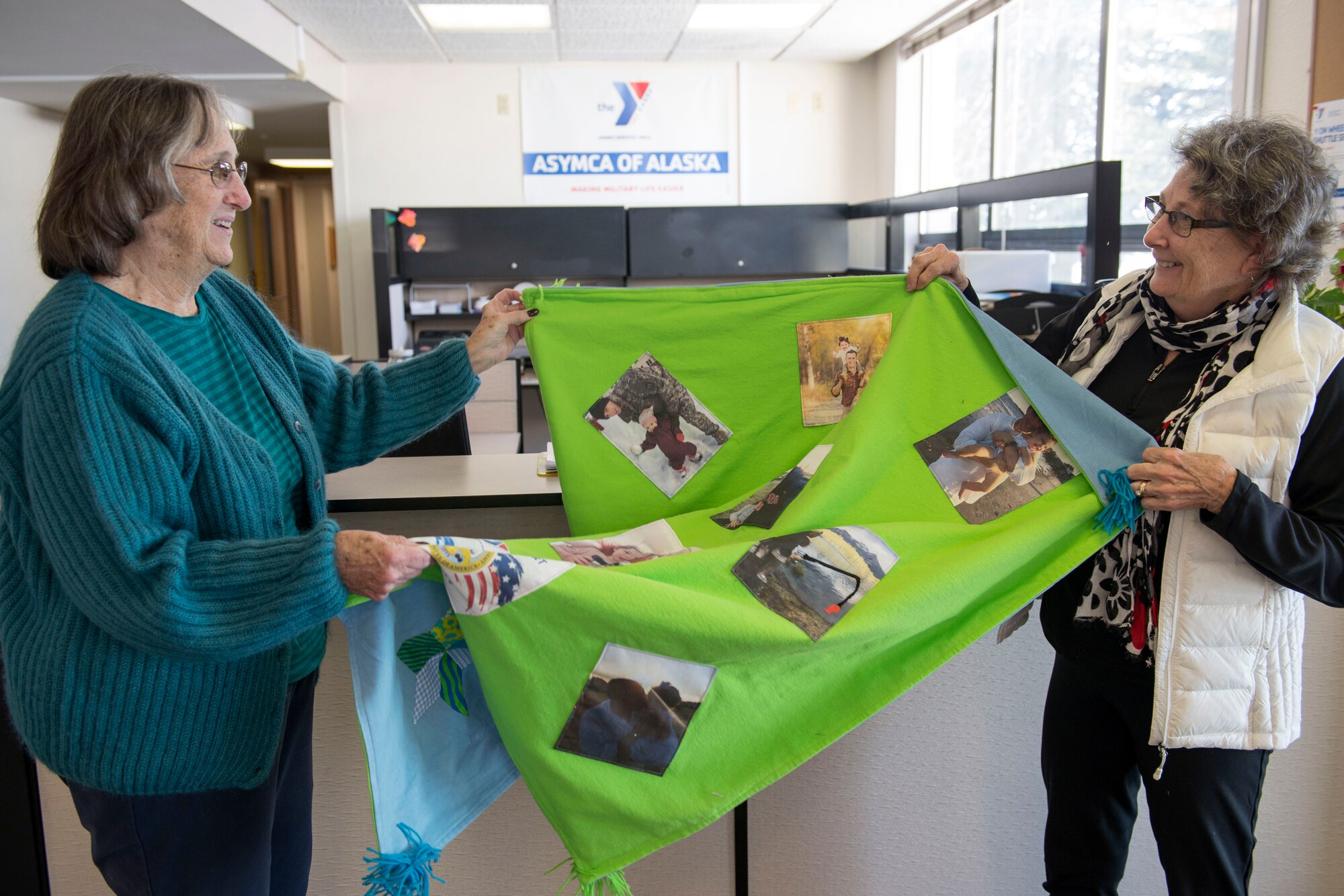 Diane Miller, Quilts from the Heart coordinator and Vee Anderson, a Quilts from the Heart volunteer, unveil a newly made quilt for a recipient of Operation Kid Comfort at the Armed Services YMCA of Alaska Joint Base Elmendorf-Richardson branch, March 26, 2018. Program volunteers at the ASYMCA of Alaska coordinate with local quilting groups to create custom-made photo-transfer quilts and pillow cases for the children of deployed active-duty service members.