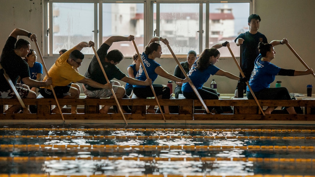 People sitting in row on crates beside a pool hold paddles in the water.