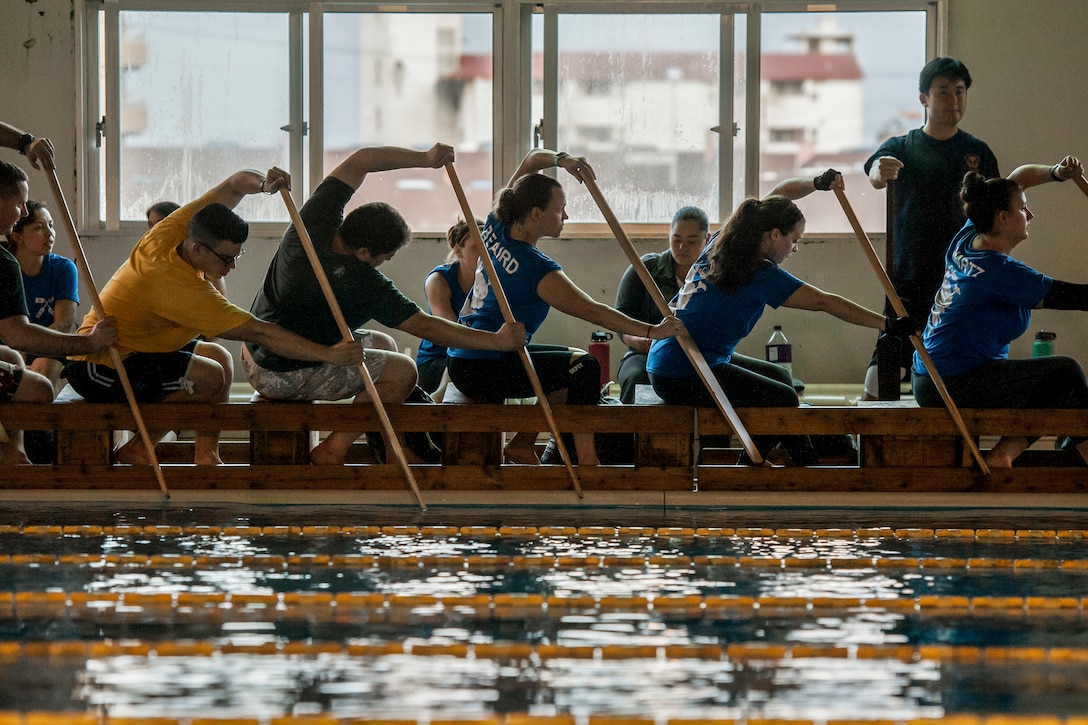 People sitting in row on crates beside a pool hold paddles in the water.