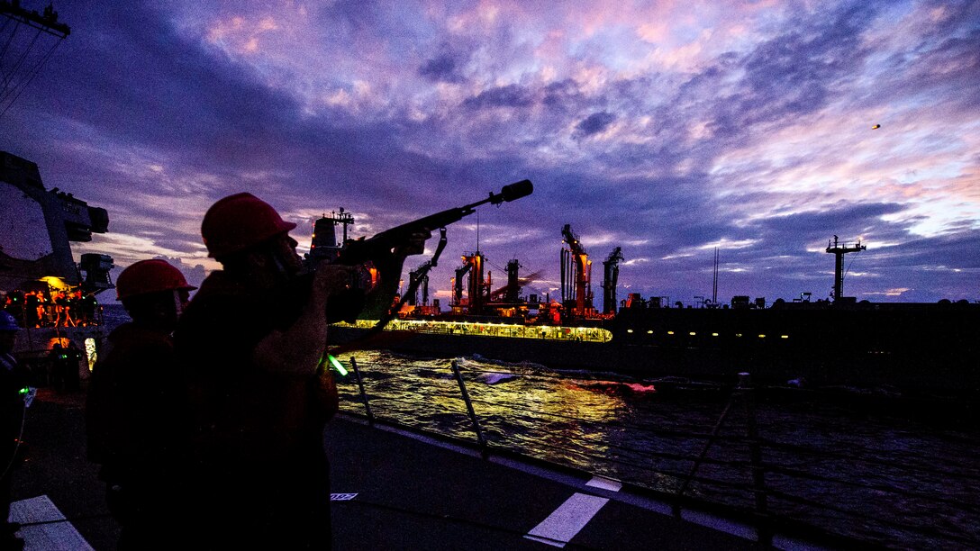 A sailor, shown in silhouette, aims a rifle at an illuminated ship traveling nearby, against a purple sky.