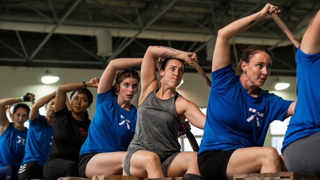 A row of women sitting on crates hold paddles and simulate rowing.