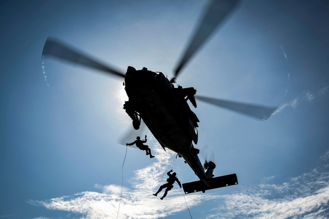 Two sailors, shown in silhouette, rappel out of a helicopter on ropes.