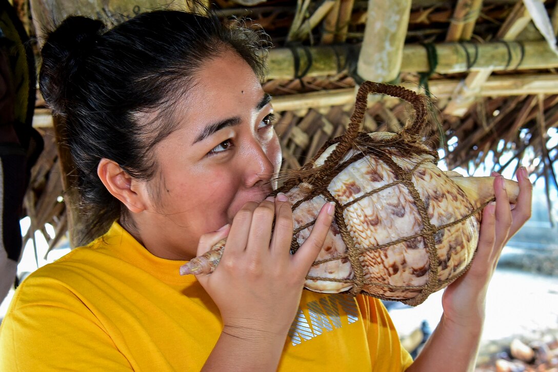 A sailor blows on a large conch under a thatched roof on a beach.