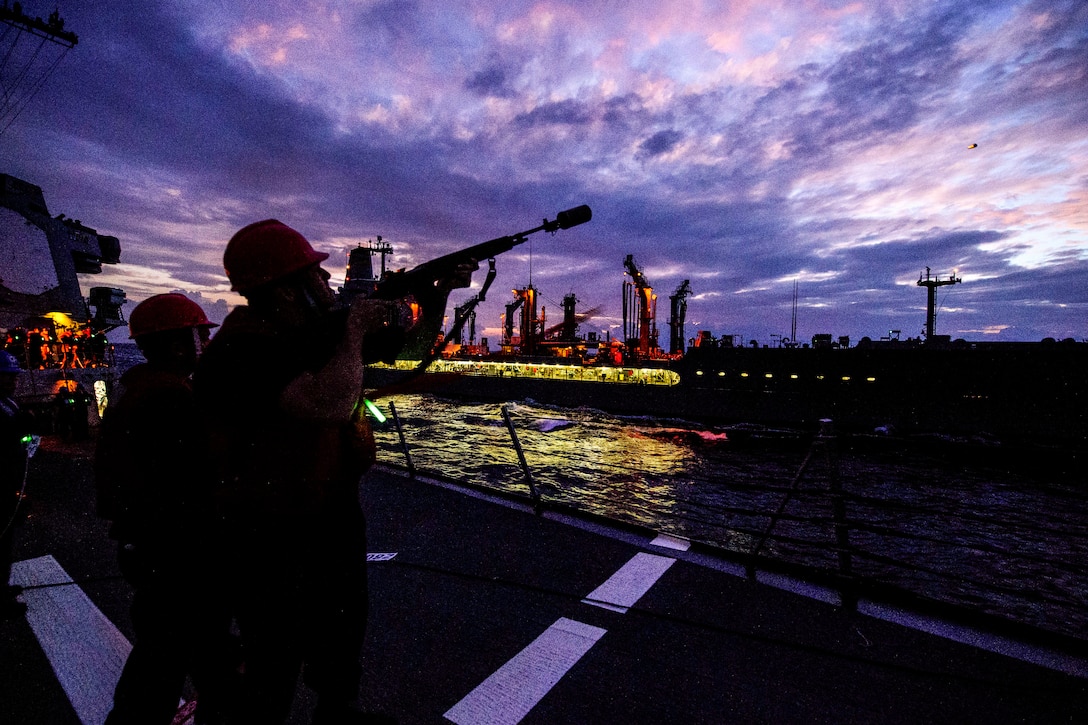 A sailor, shown in silhouette, aims a rifle at an illuminated ship traveling nearby, against a purple sky.