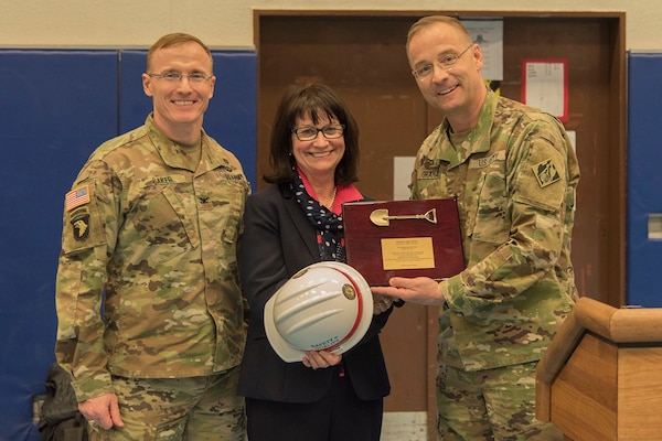 Two men and one woman stand and smile for the camera. The woman is holding a hard hat and a plaque.