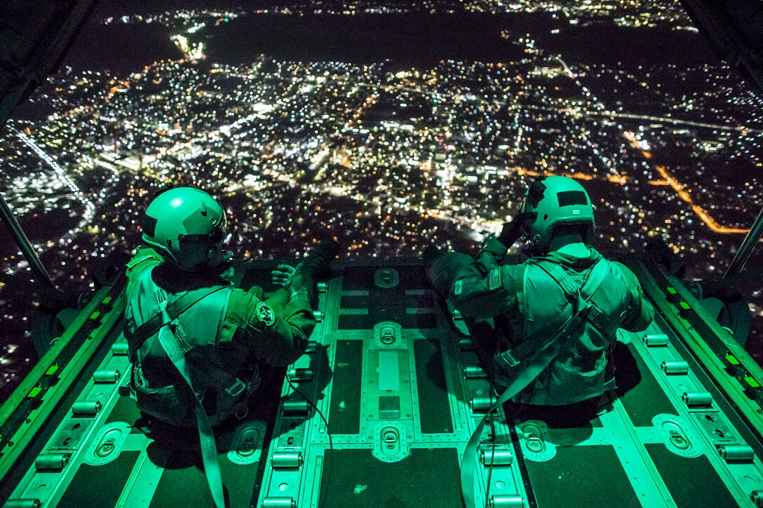 Two airmen sit on the ramp of an aircraft flying at night over a city.