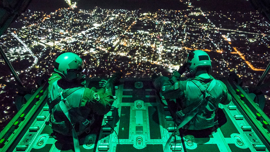 Two airmen sit on the ramp of an aircraft flying at night over a city.