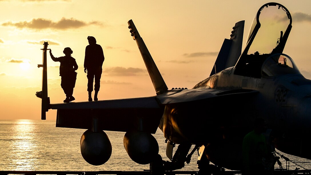 Two sailors stand on an aircraft on a ship's deck and watch the sunset.
