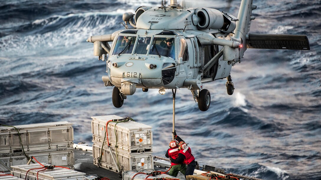 Two sailors help a helicopter offload ammo while at sea.