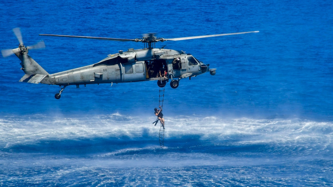 A sailor climbs a rope ladder up to a helicopter hovering over turquoise water.