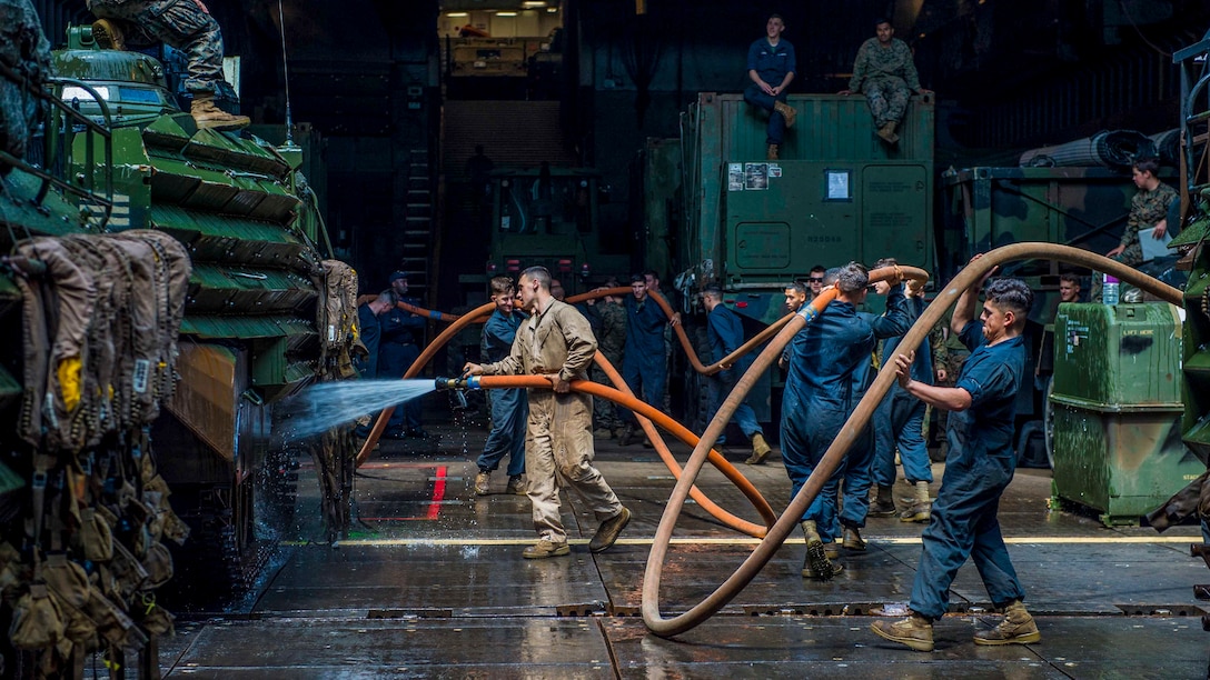 Marines maneuver large hoses in a dark, damp space while washing vehicles.