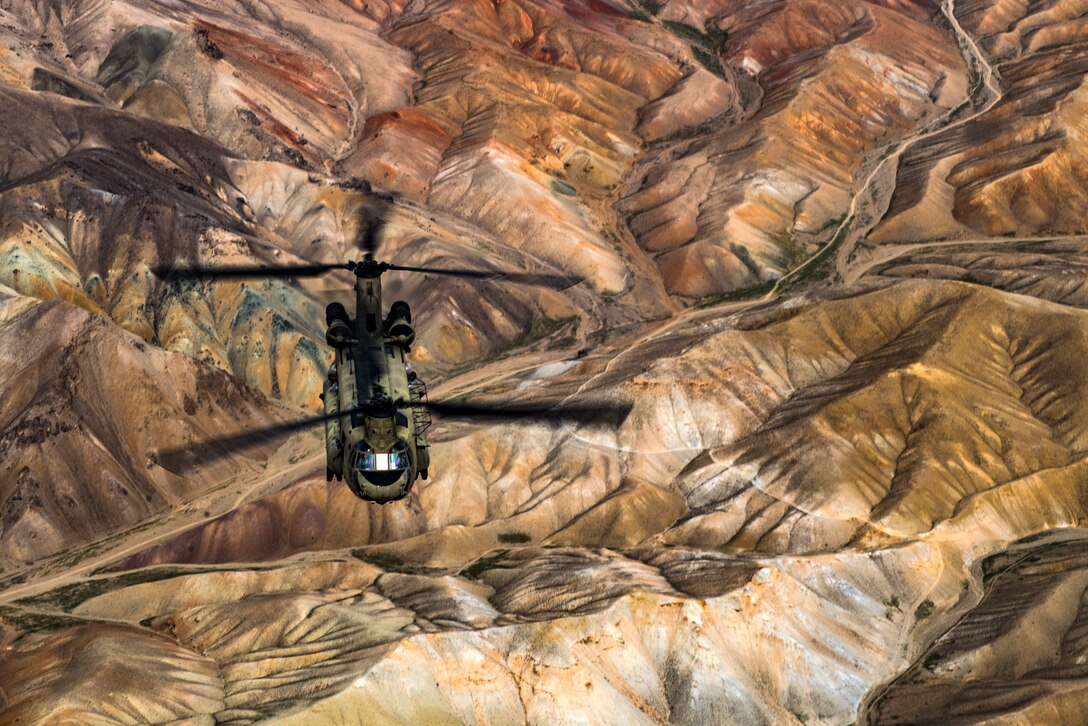 A helicopter, shown from above, flies over hilly, brown terrain.