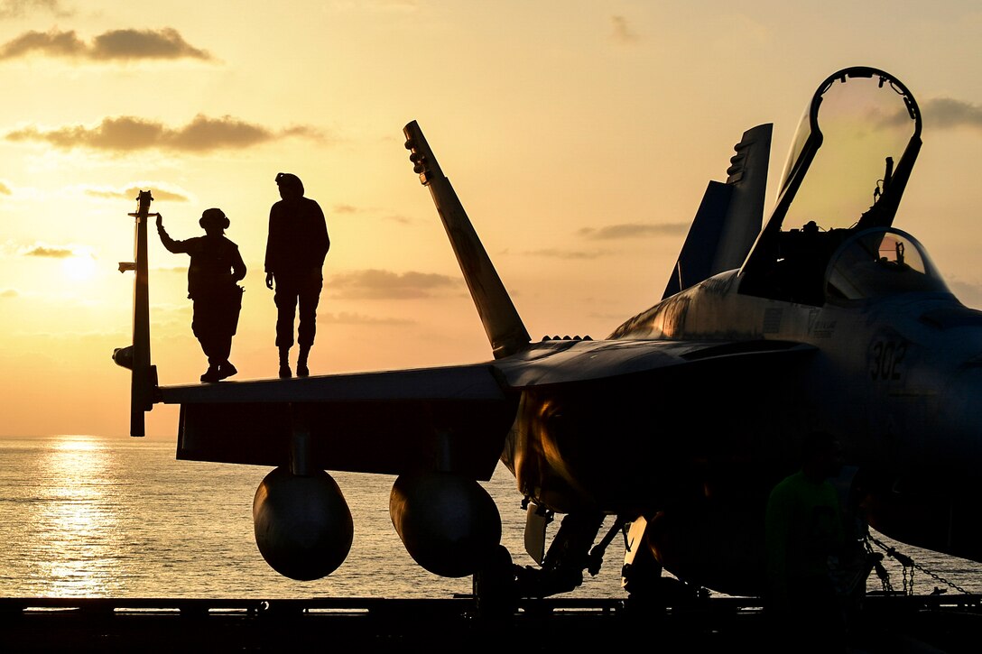 Sailors watch the sunset on an aircraft on a ship's flight deck.