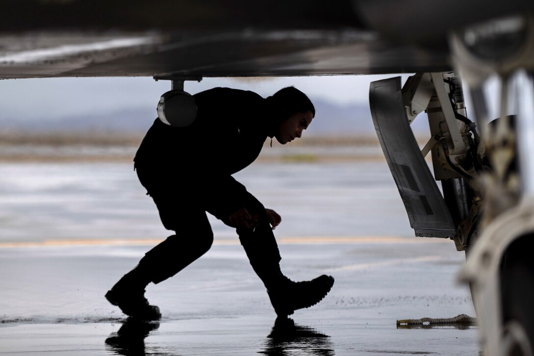 An airman crouches while walking beneath an aircraft wing on a wet flightline.