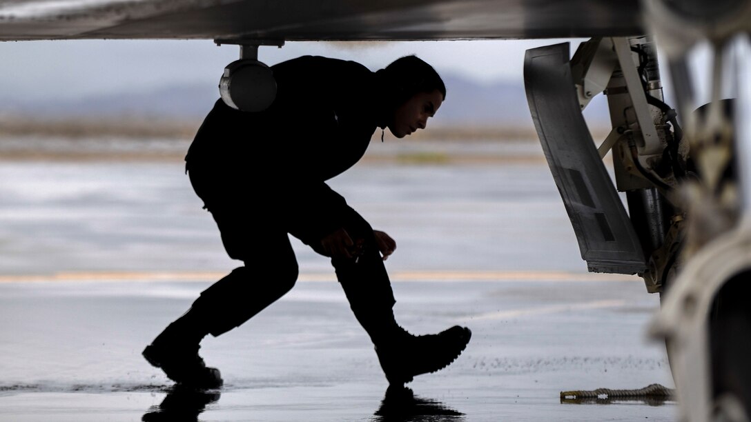 An airman crouches while walking beneath an aircraft wing on a wet flightline.