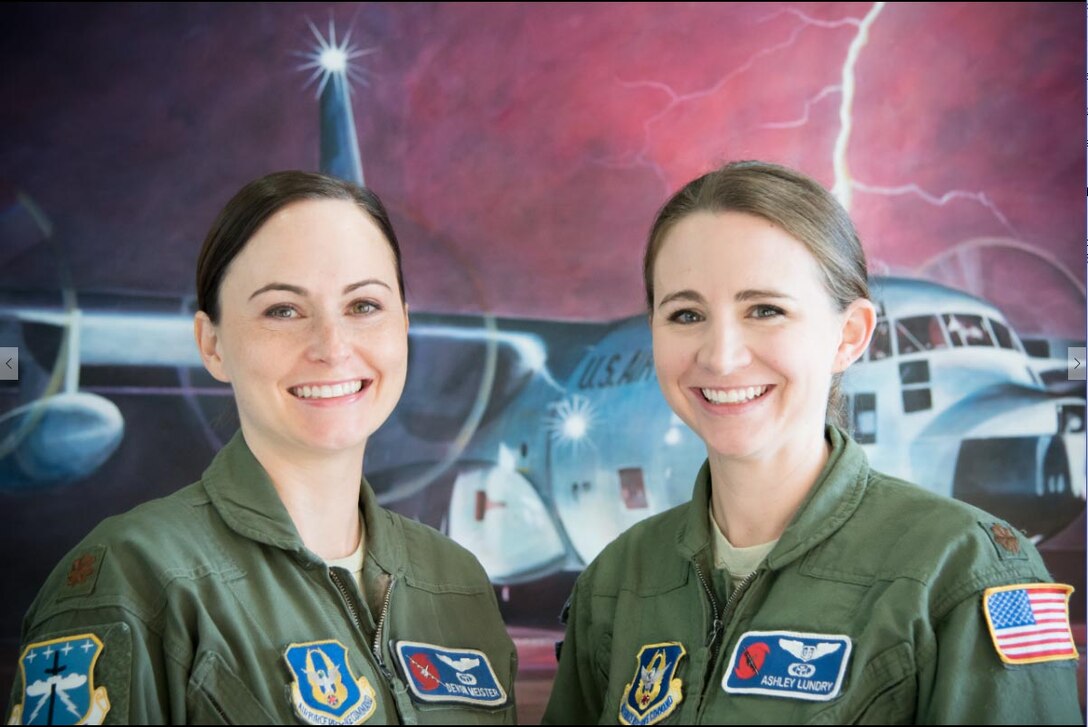 Two airmen smile at the camera with a lightning over aircraft background.