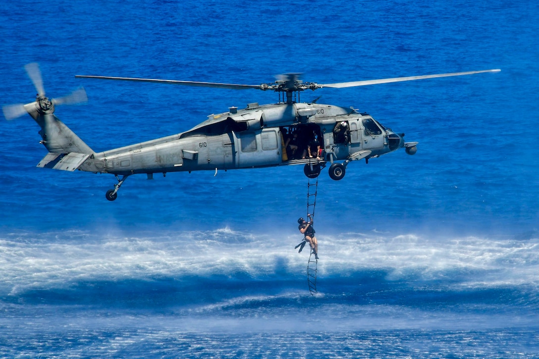 A sailor climbs a rope ladder up to a helicopter hovering over turquoise water.