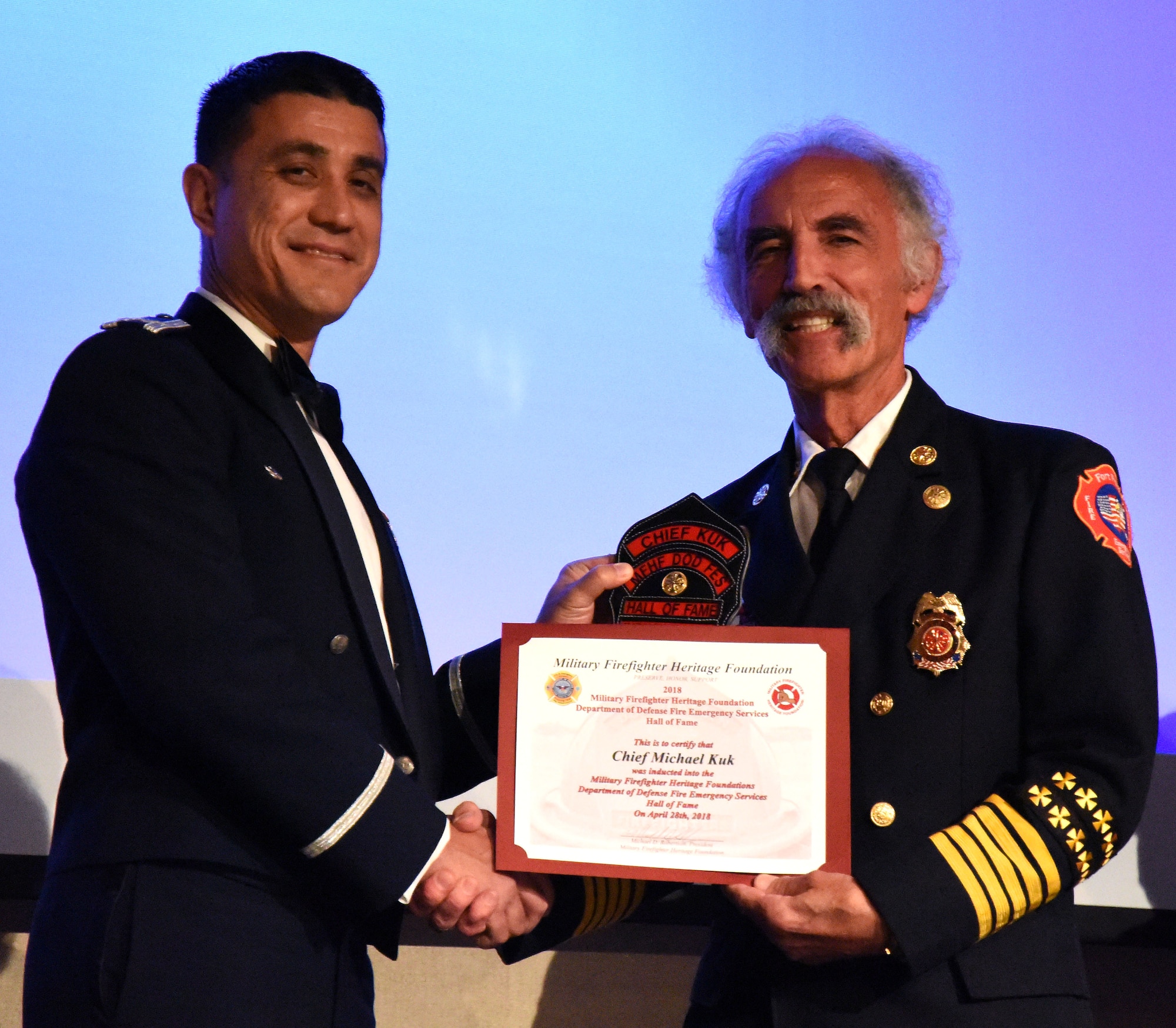 U.S. Air Force Col. Ricky Mills, 17th Training Wing commander, presents Chief Michael Kuk, firefighter Hall of Fame inductee, with a new clip for the front of his helmet as congratulations during the 16th Annual Firefighter Ball at the McNease Convention Center in San Angelo Texas, April 28, 2018. Kuk began his career as a firefighter in the U.S. Army from 1969-1975, he is now a military firefighting historian. (U.S. Air Force photo by Airman 1st Class Seraiah Hines/Released)