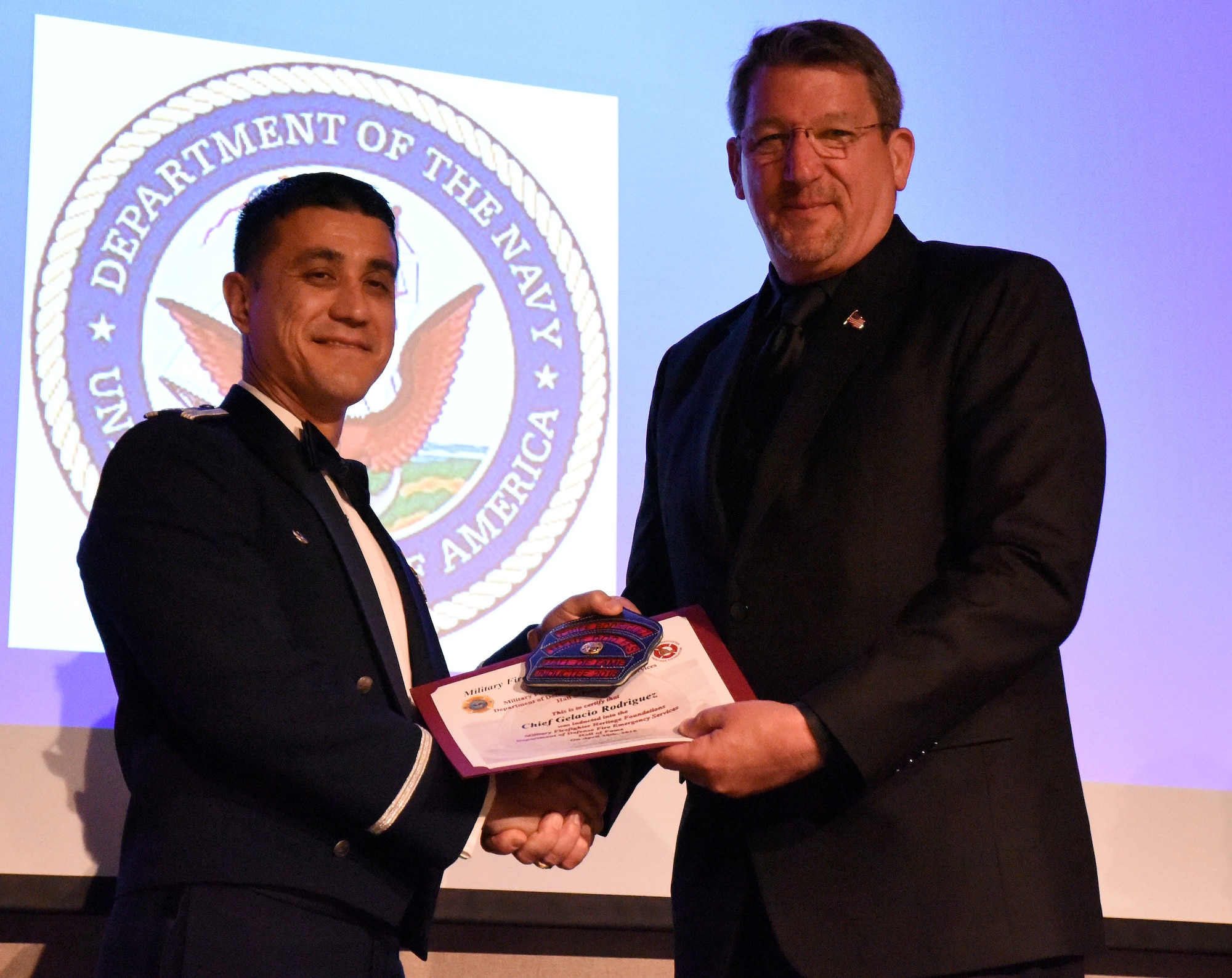U.S. Air Force Col. Ricky Mills, 17th Training Wing commander, presents Chief Gene Rausch, guest of honor, with a clip for a firefighter helmet as congratulations during the 16th Annual Firefighter Ball at the McNease Convention Center, San Angelo, Texas, April 28, 2018. The hall of fame inductee award is intended for Chief Gelacio Rodriquez, firefighter hall of fame inductee, who could not attend the ball. (U.S. Air Force photo by Airman 1st Class Seraiah Hines/Released)