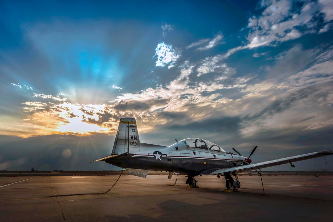 A small military aircraft sits on the tarmac as the sun peeks through the clouds.