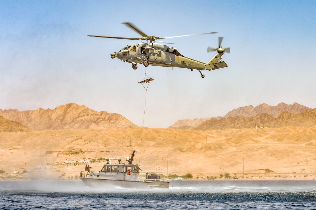 A stretcher hangs on a rope from a helicopter hovering over a boat in the water, with brown hills in the background.