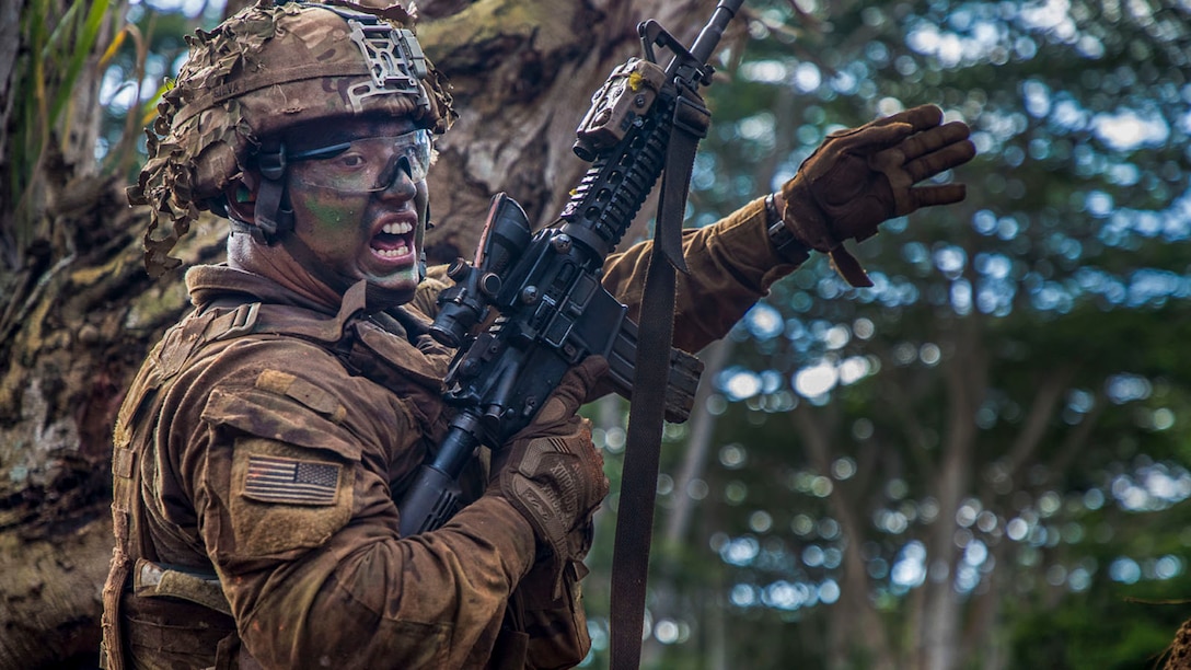 A soldier in camouflage holding a weapon waves his arm and shouts in a wooded area.