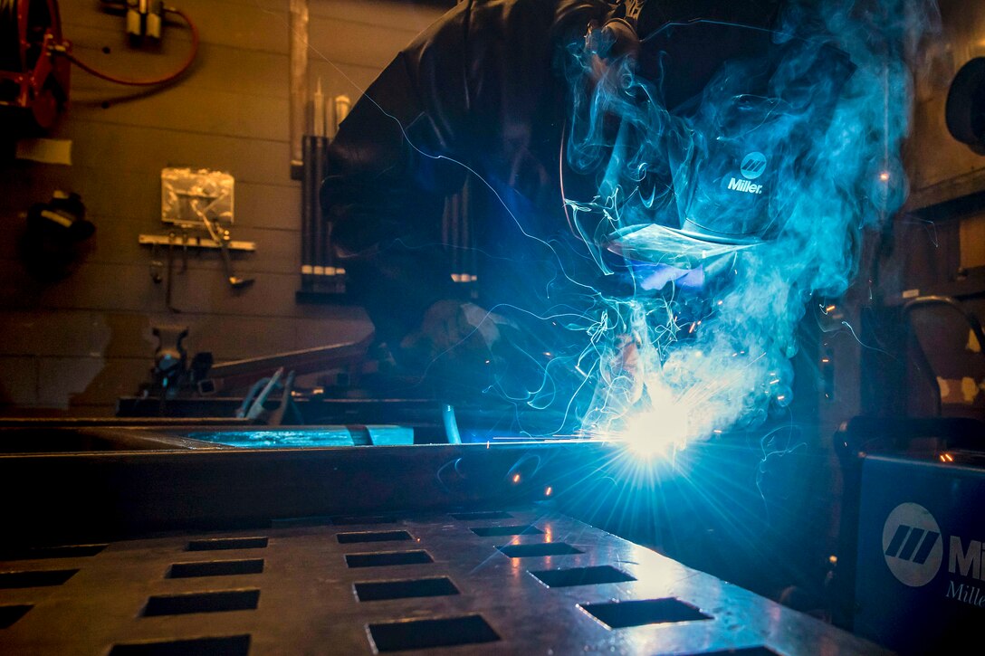 Blue smoke wafts upward in a room as an airman in a protective helmet does welding-type work.