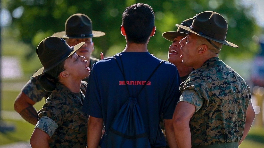 Drill sergeants stand in circle around a Marine Corps recruit, shown from behind, and shout at him.