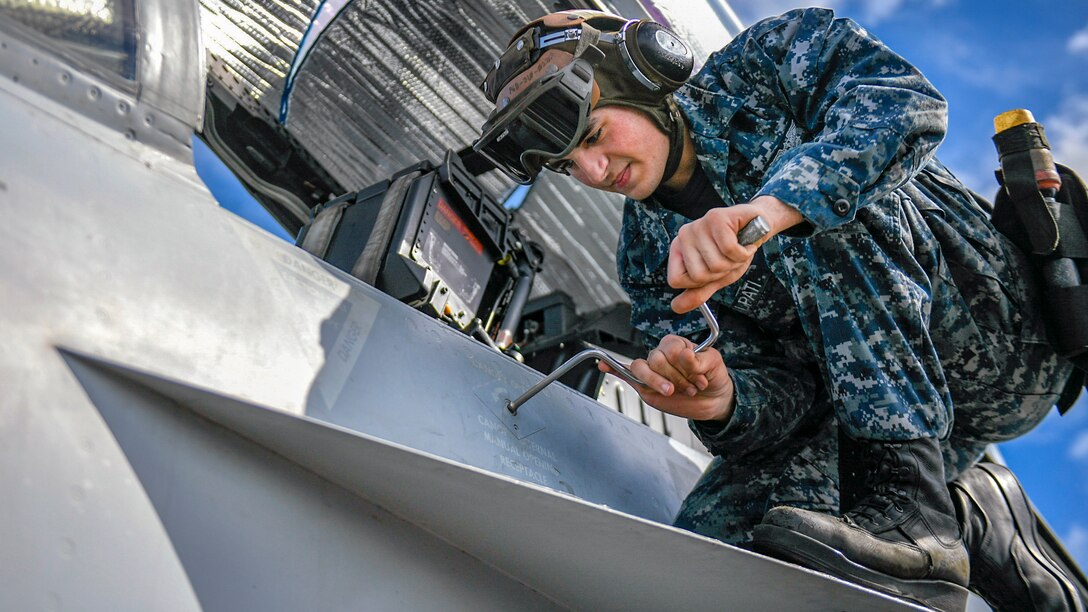A sailor manipulates a tool while kneeling on an aircraft wing and working on the aircraft.