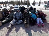Senior Airman Elliott Bull (center left) and local children from Shungnak, Alaska, hunker down for shelter amidst strong winds while waiting for a helicopter to arrive
