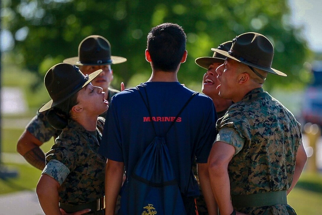 Drill sergeants stand in circle around a Marine Corps recruit, shown from behind, and shout at him.