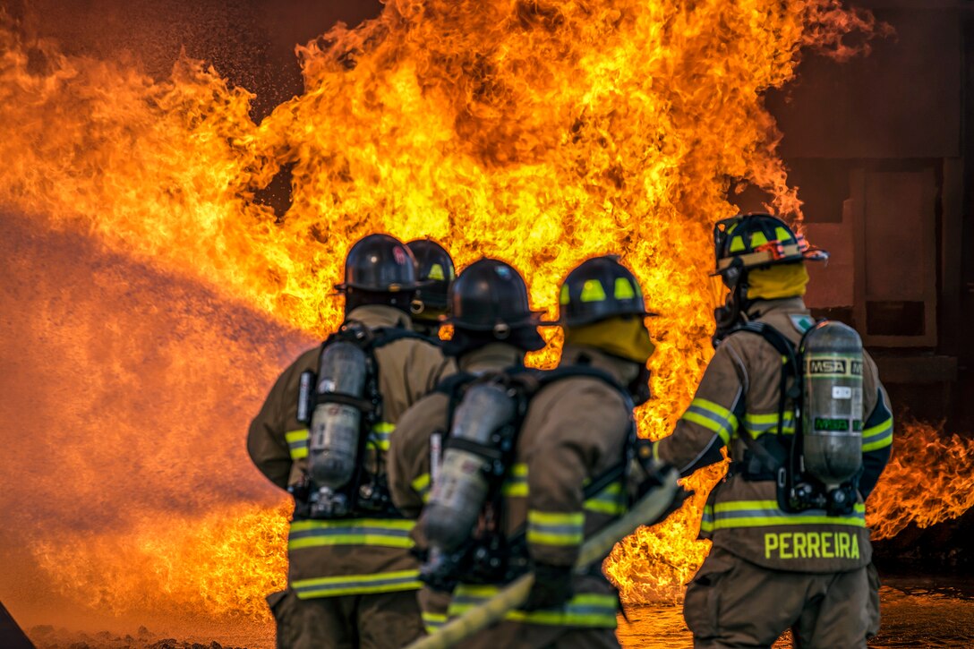 Five firefighters, shown from behind, use a hose to spray water on a fire.