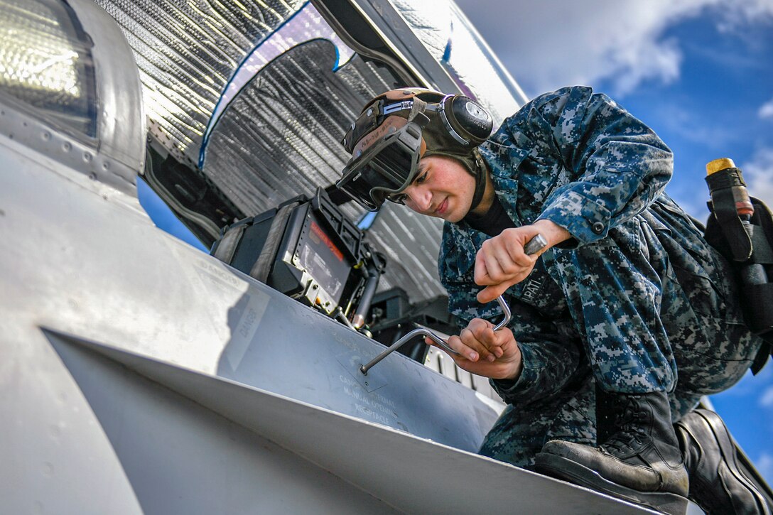 A sailor manipulates a tool while kneeling on an aircraft wing and working on the aircraft.