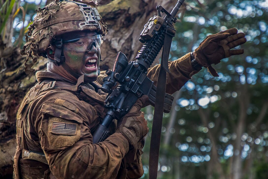 A soldier in camouflage face paint holds a weapon in one arm and waves the other while shouting in a wooded area.