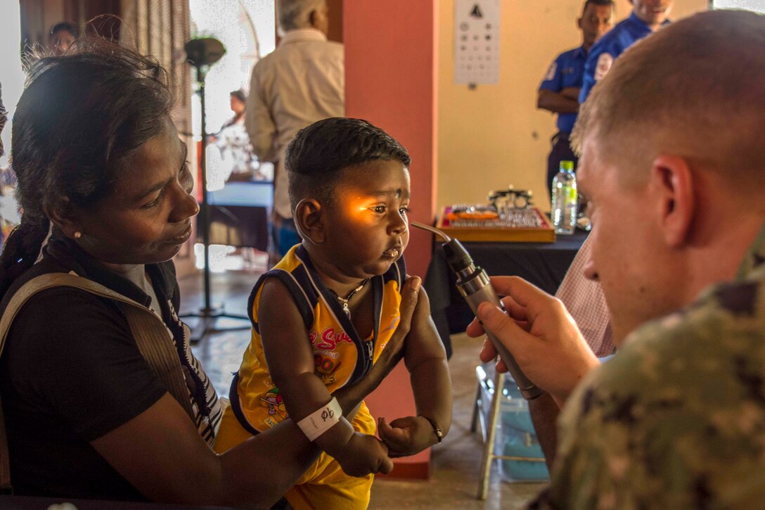 A sailor uses a light to examine the eyes of a toddler who is being held by a woman.