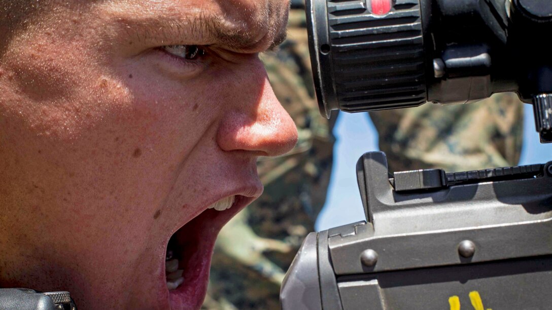 A Marine, shown in close-up profile, shouts with his mouth wide open, while behind a weapon.