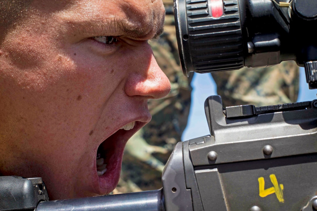 A Marine, shown in close-up profile, shouts with his mouth wide open, while behind a weapon.