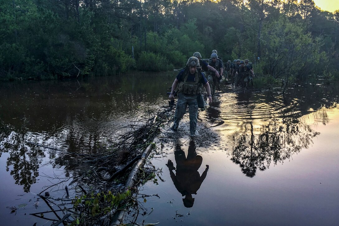 Soldiers wade through knee-deep waters, which show their reflections and those of the trees around them.