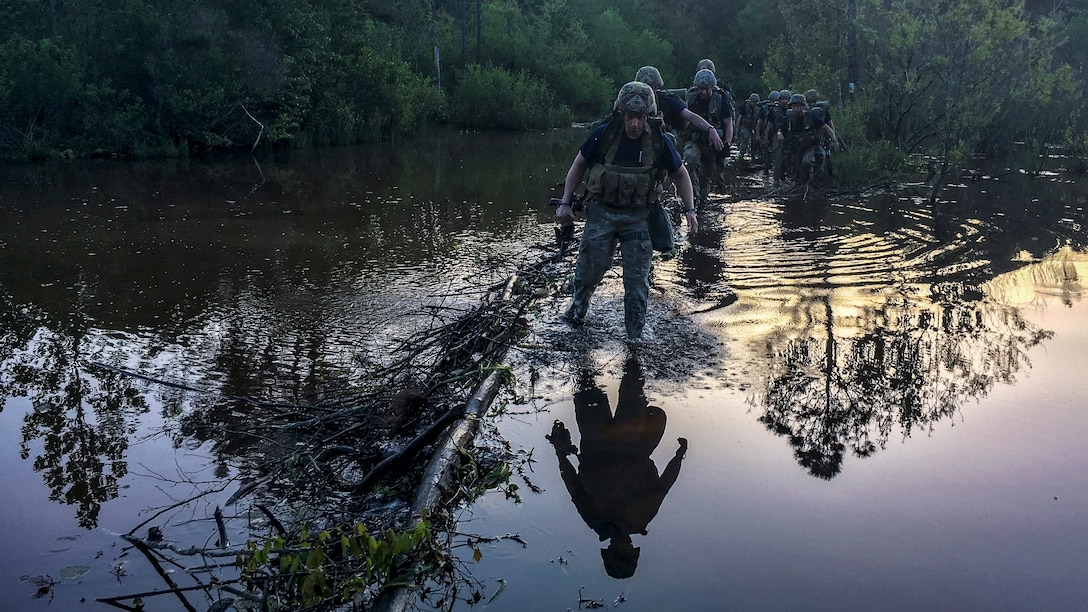 Soldiers wade through knee-deep waters, which show their reflections and those of the trees around them.