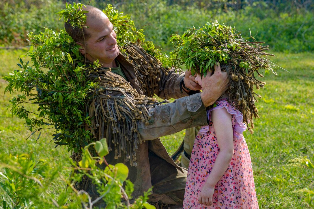 A kneeling Marine puts a cap of foliage on a child's head in a green field.