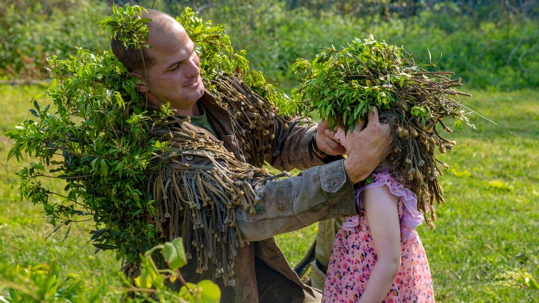 A kneeling Marine puts a cap of foliage on a child's head in a green field.