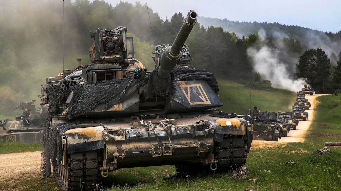 A tank leads a long line of tanks driving on a dusty dirt road over green terrain.