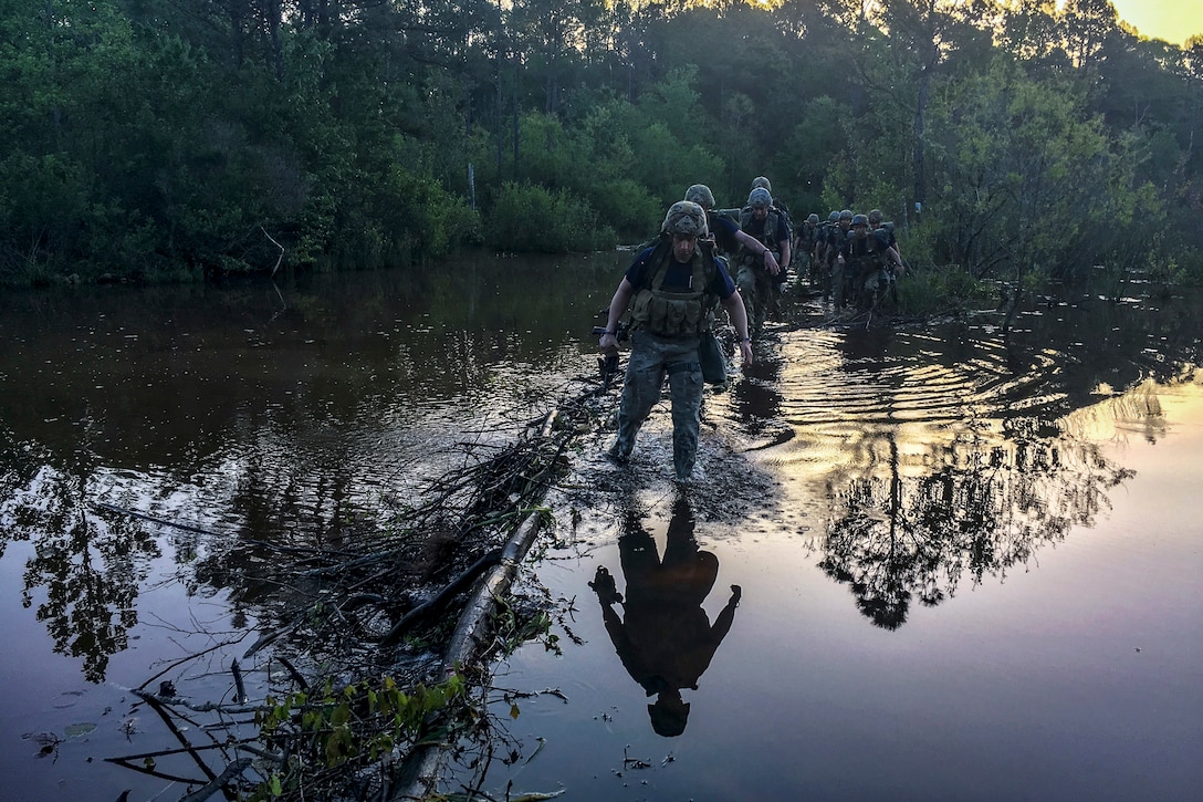 Soldiers wade through knee-deep waters, which show their reflections and those of the trees around them.