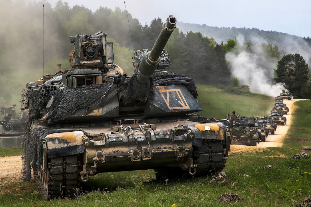 A tank leads a long line of tanks driving on a dusty dirt road over green terrain.